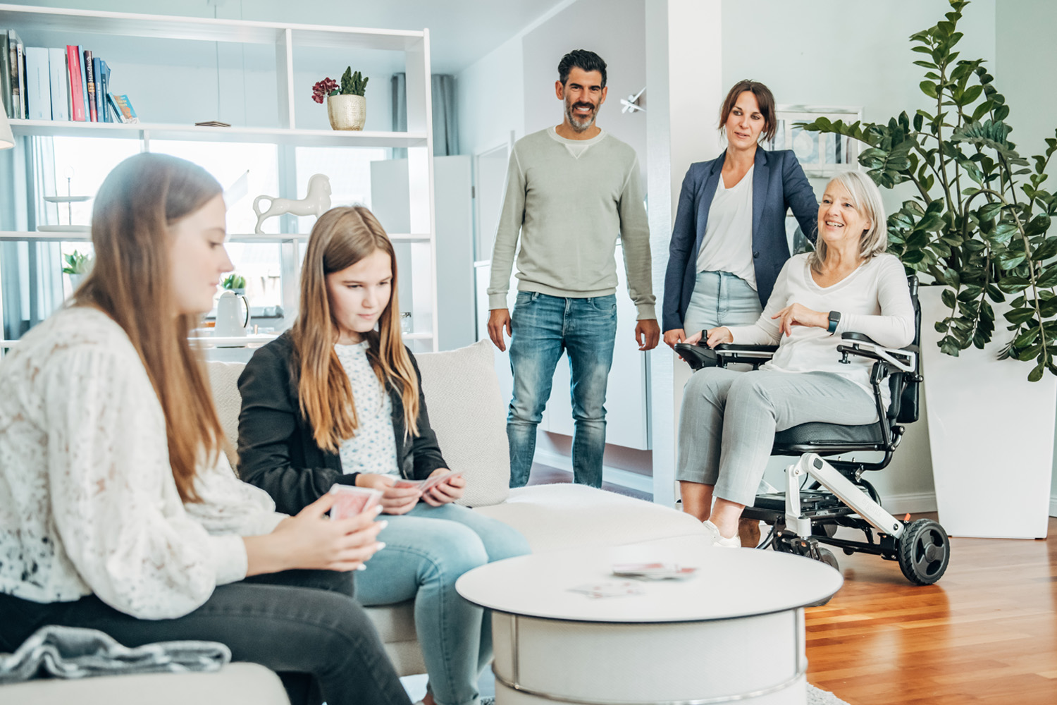 Das Bild zeigt eine Familie in der Wohnung. Die Kinder spielen im Vordergrund und die Oma im Rollstuhl schaut mit den Eltern zu